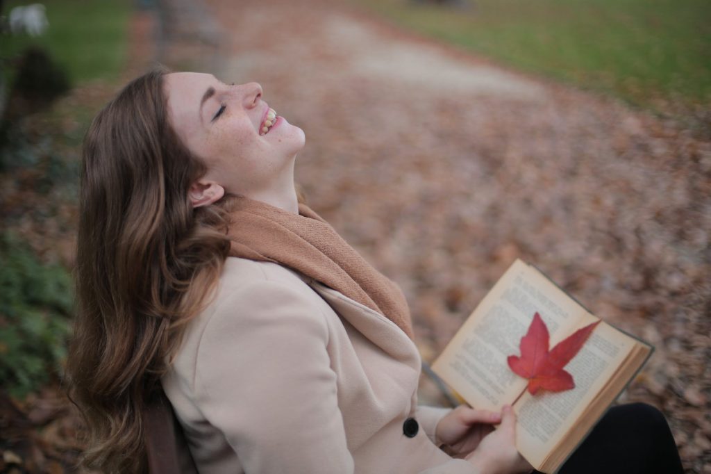 cheerful young woman with red leaf enjoying life and weather while reading book in autumn park