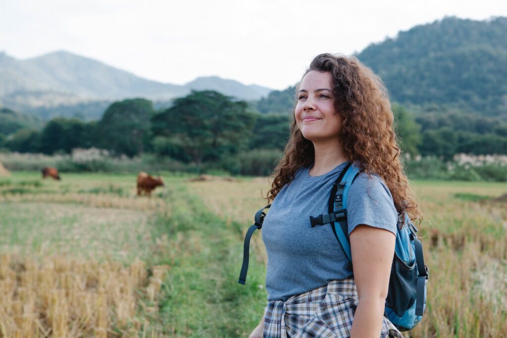 positive female traveler with curly hair standing on green field against hills