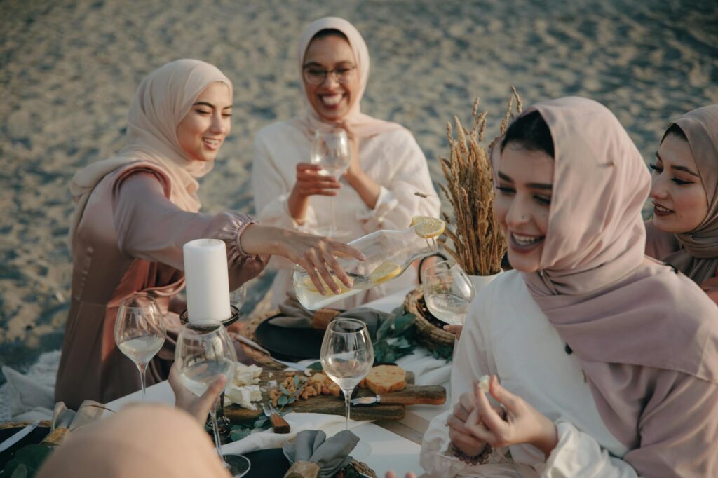 group of women wearing hijab sitting on beach sand drinking and celebrating