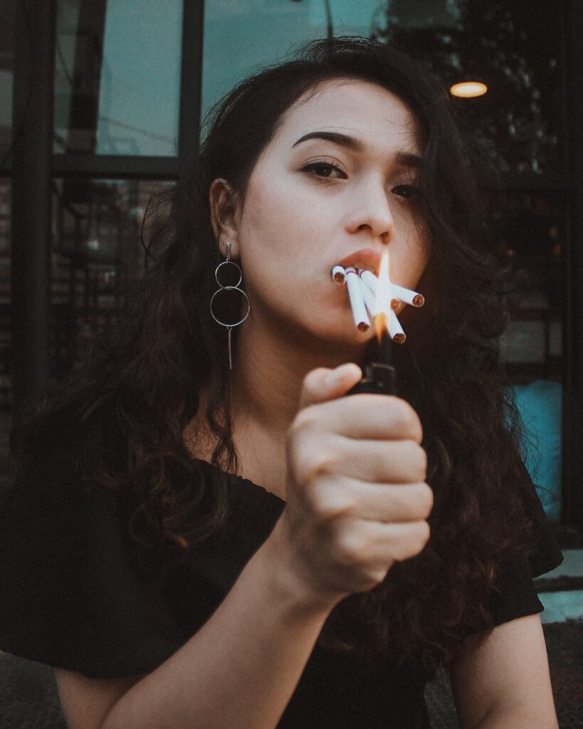 pensive young ethnic female smoker with lighter and cigarettes in mouth